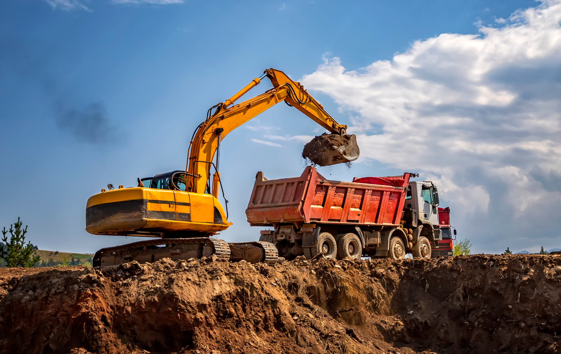 Yellow excavator loading dirt into a red dump truck on a construction site, blue sky.