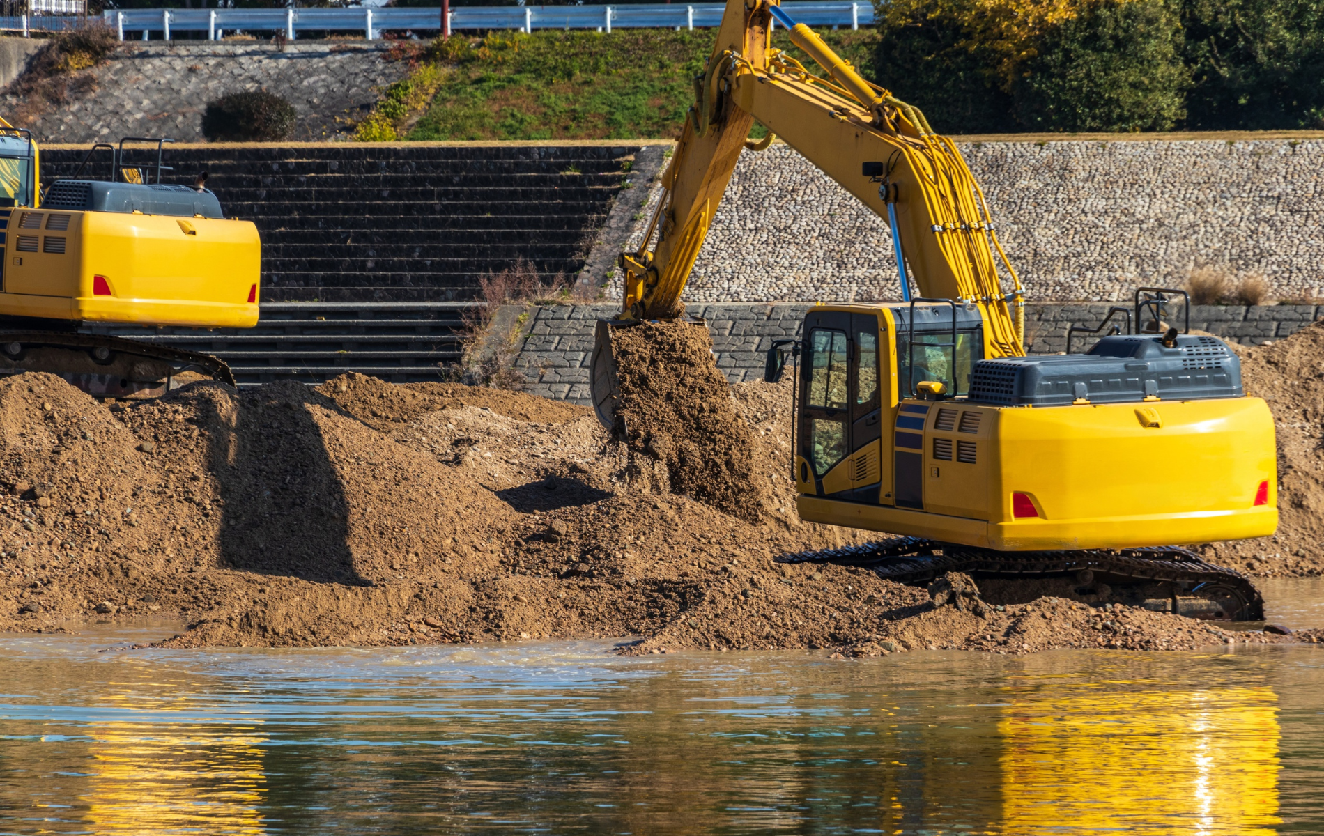 Yellow excavator digging in the water near a rocky bank.