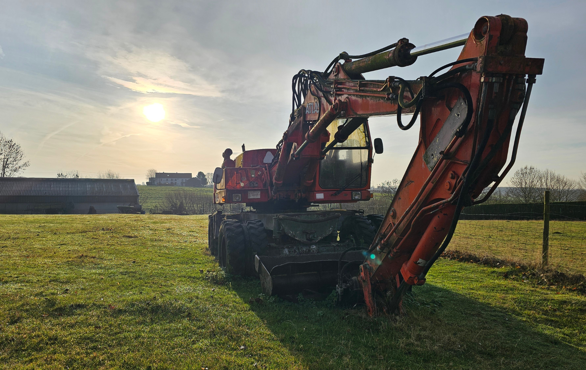Red excavator on a grassy field with a bright sun in the background.