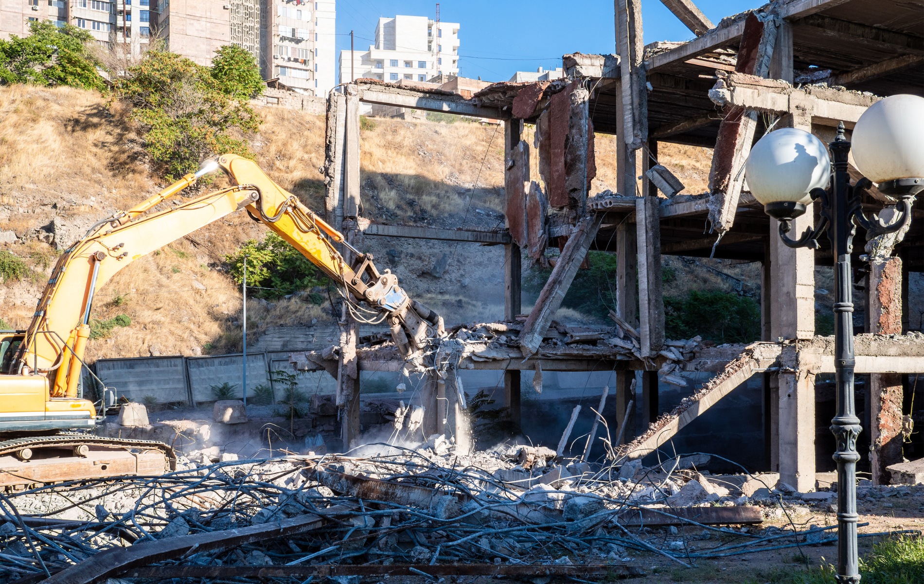 Yellow excavator demolishing a building; debris and a hillside are in the background.