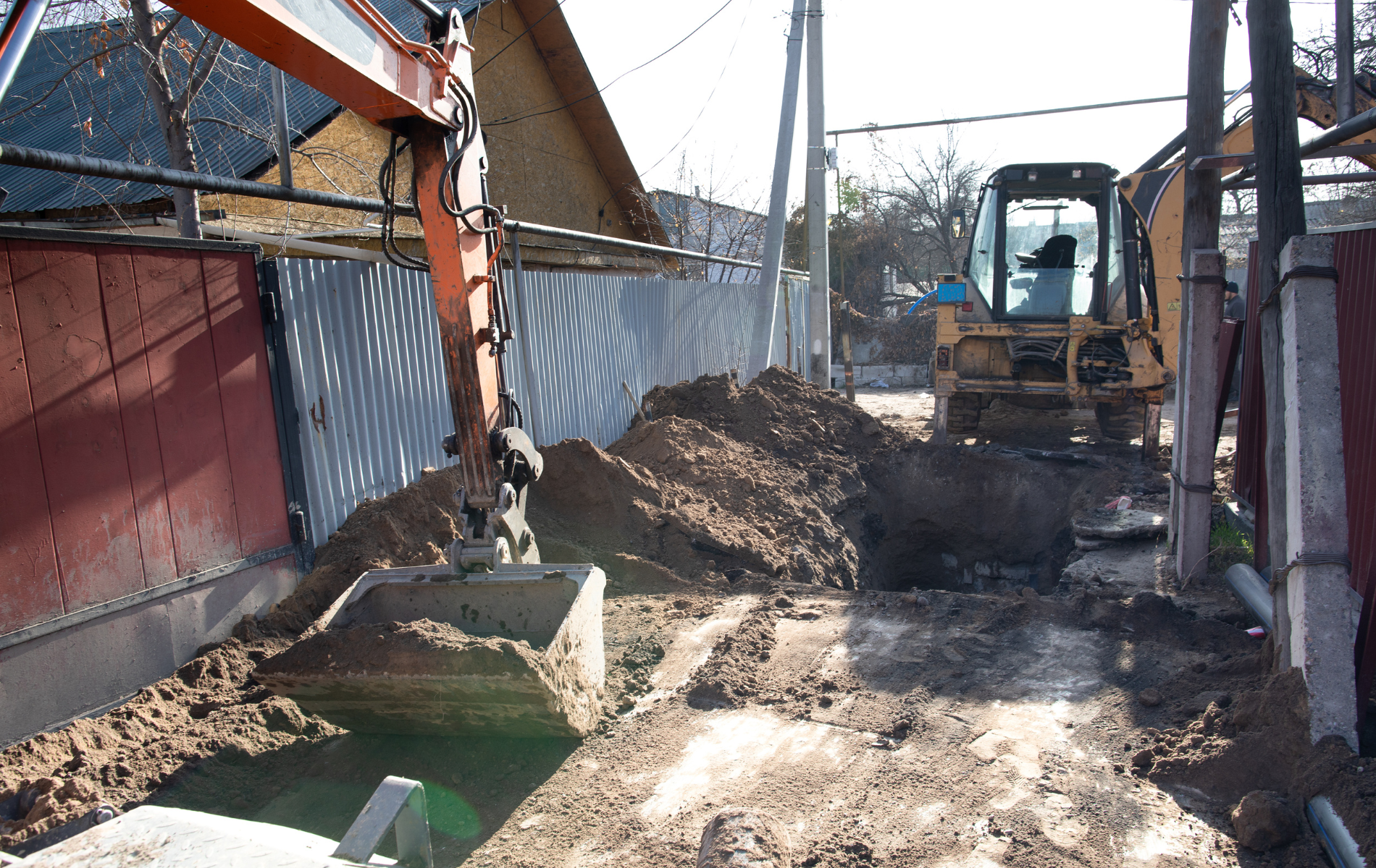 An excavator digs a trench next to a fence; a bulldozer is behind it.