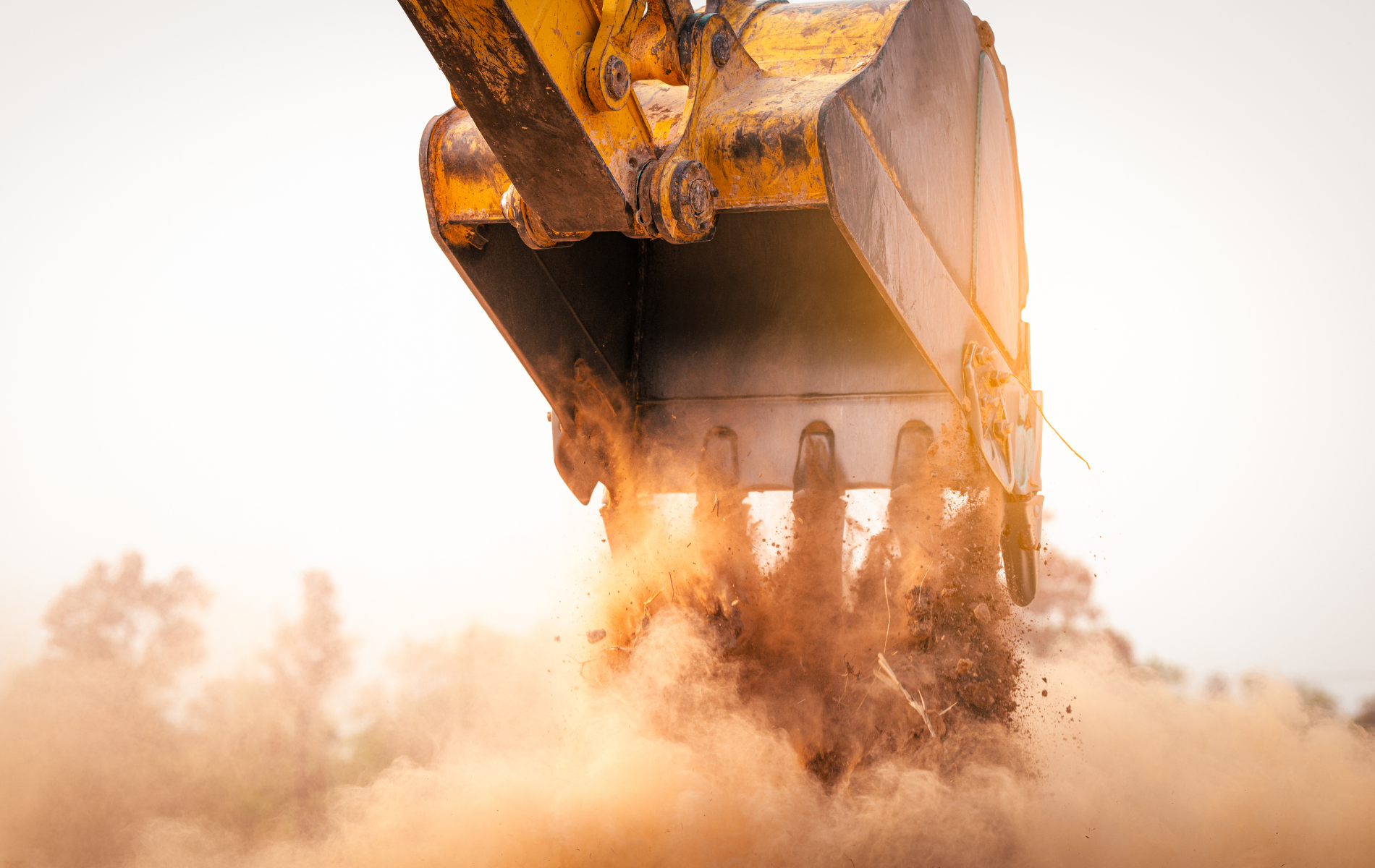 Yellow excavator bucket digging into dirt, creating a cloud of dust in the sunlight.