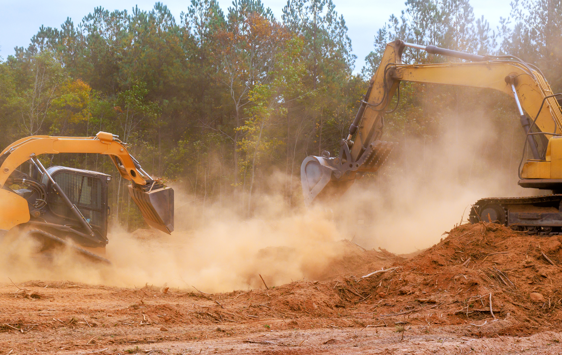 Two yellow excavators digging in dirt, creating dust. Trees in the background.