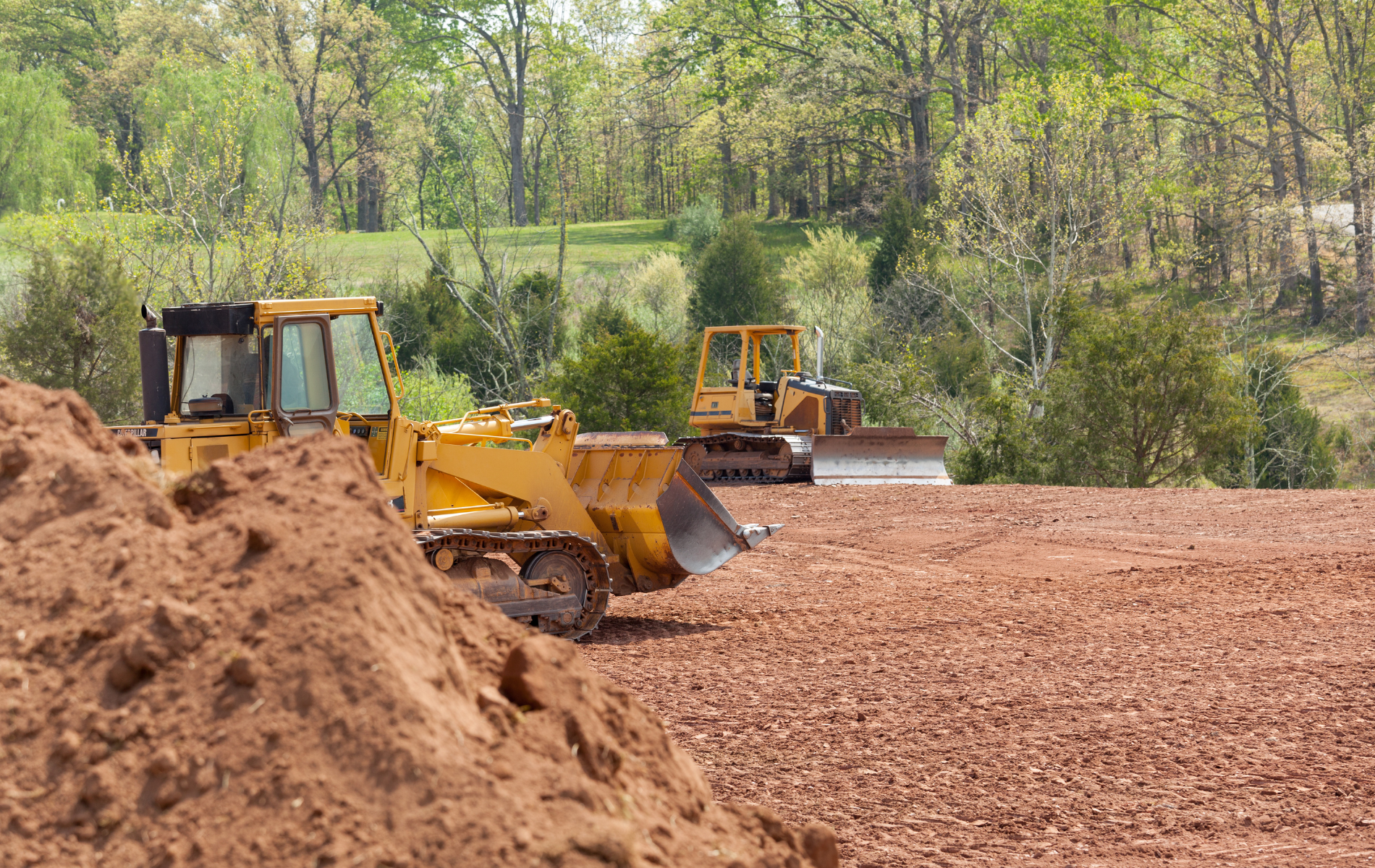 Yellow bulldozers on a reddish-brown construction site, clearing land near trees.