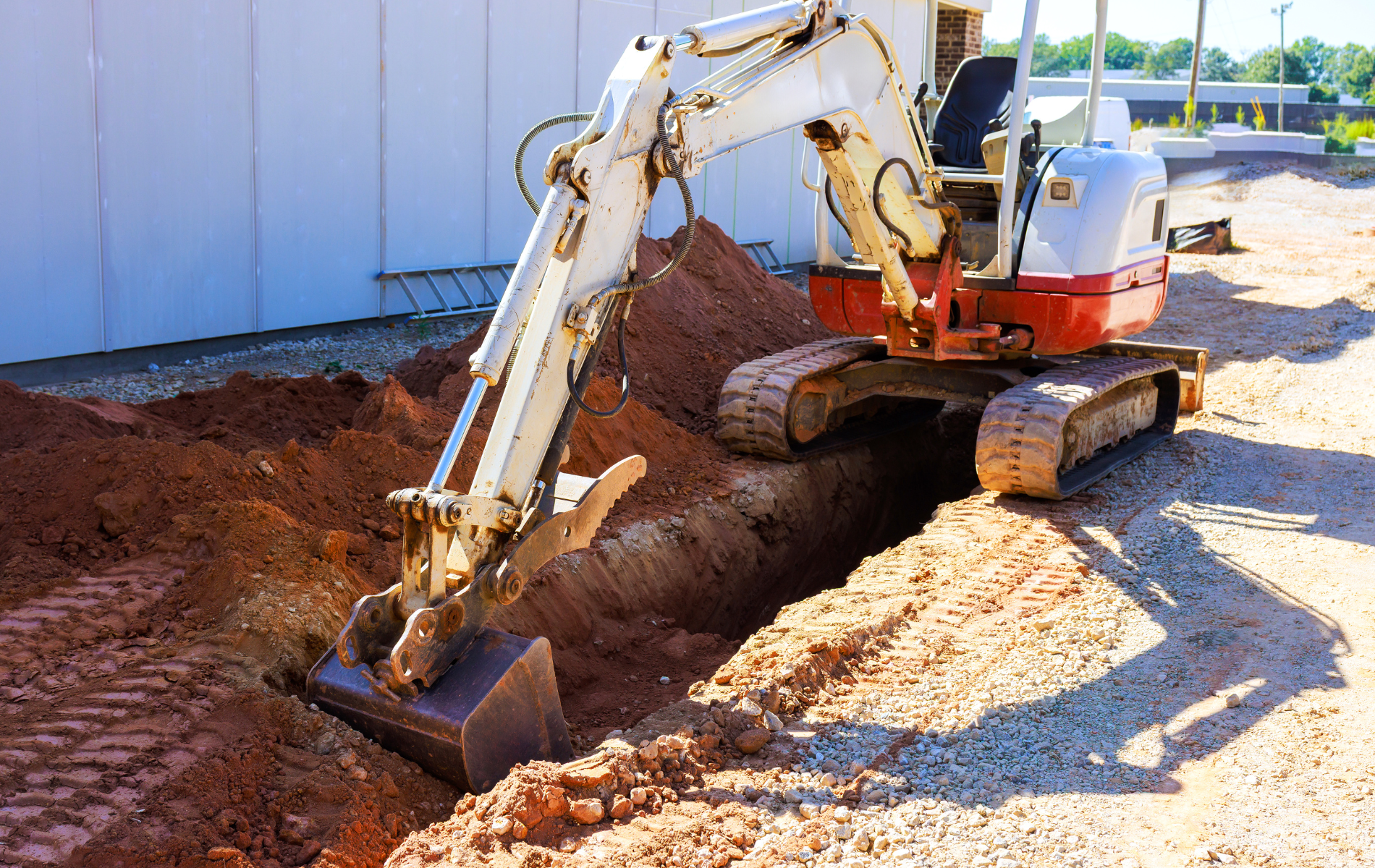 Excavator digging a trench in the dirt near a white building.