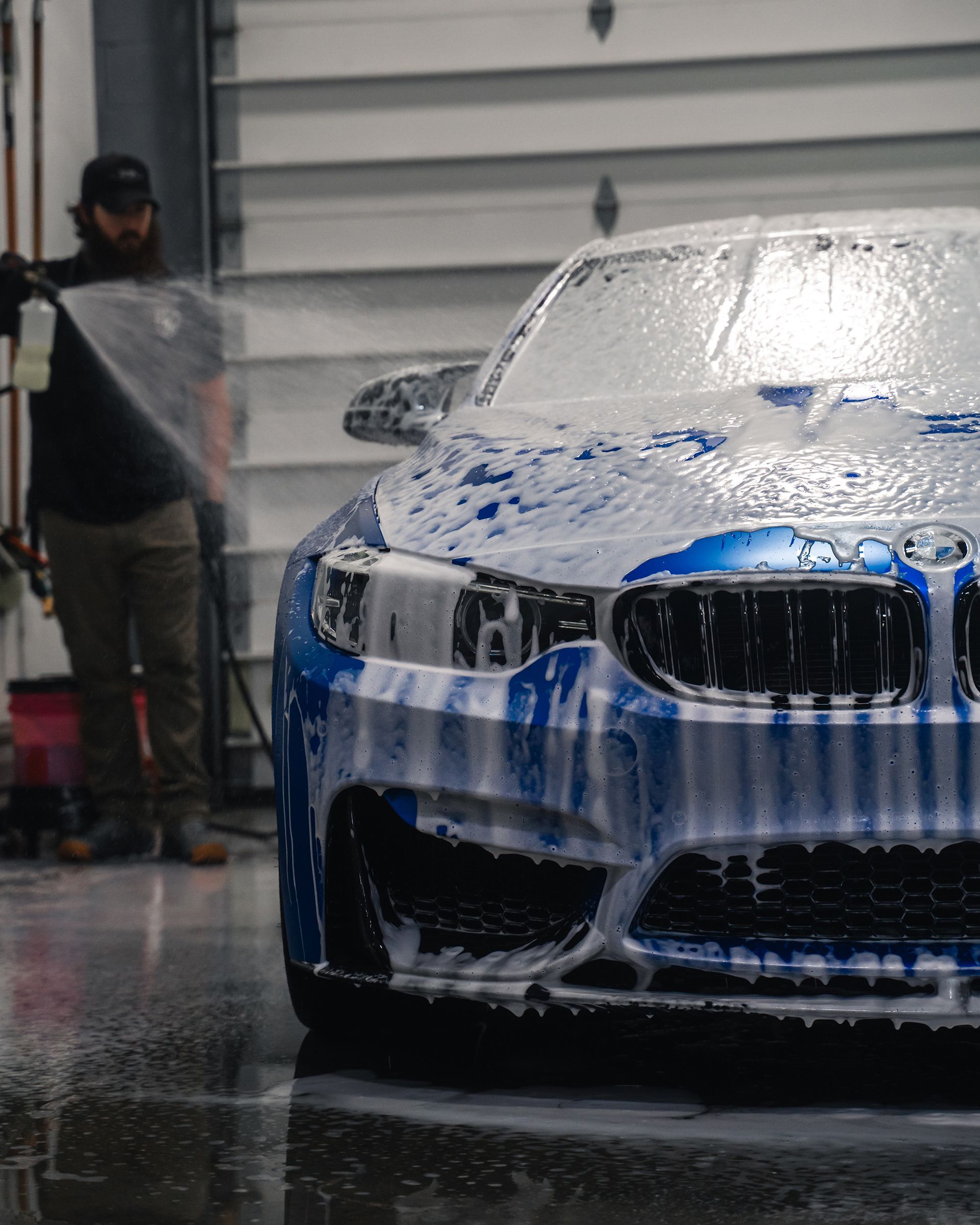 A blue BMW being foamed with soap by a person in a garage.