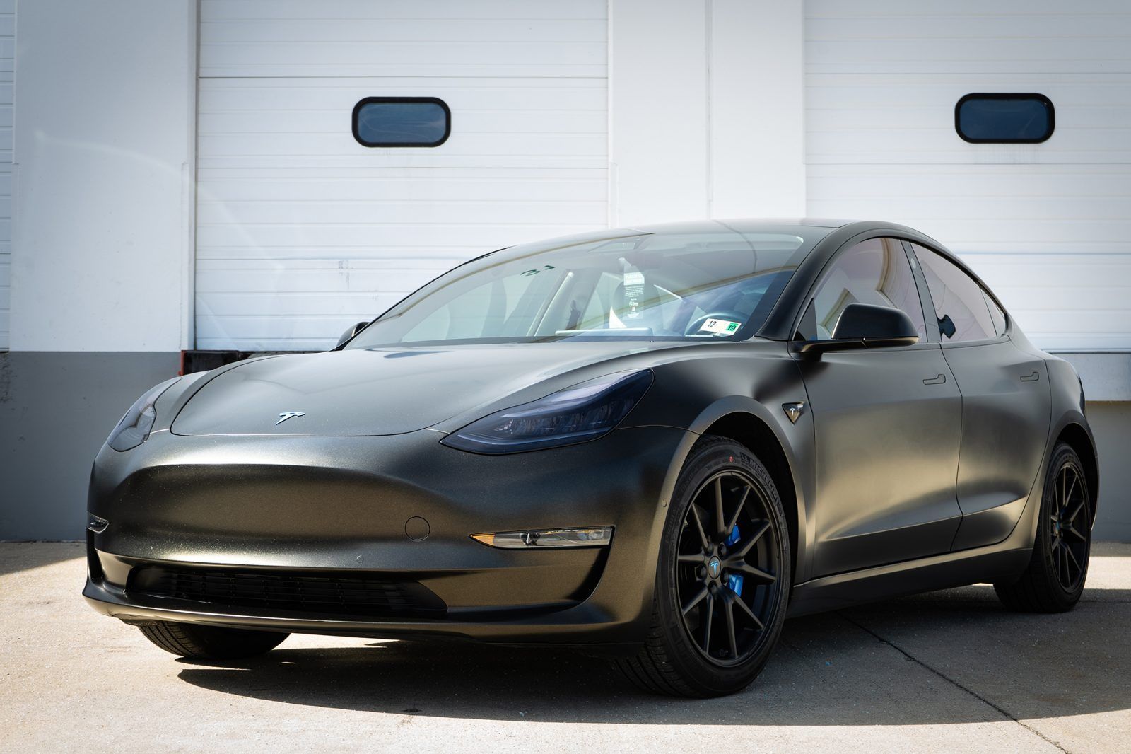 Black Tesla Model 3 parked in front of a white garage door, with tinted windows and black wheels.
