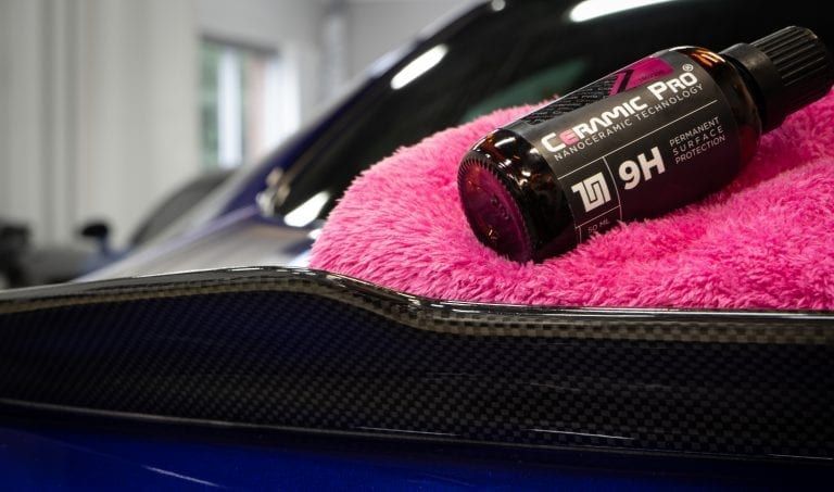 Ceramic Pro 9H coating bottle on pink microfiber towel, applied to a blue car's carbon fiber spoiler.