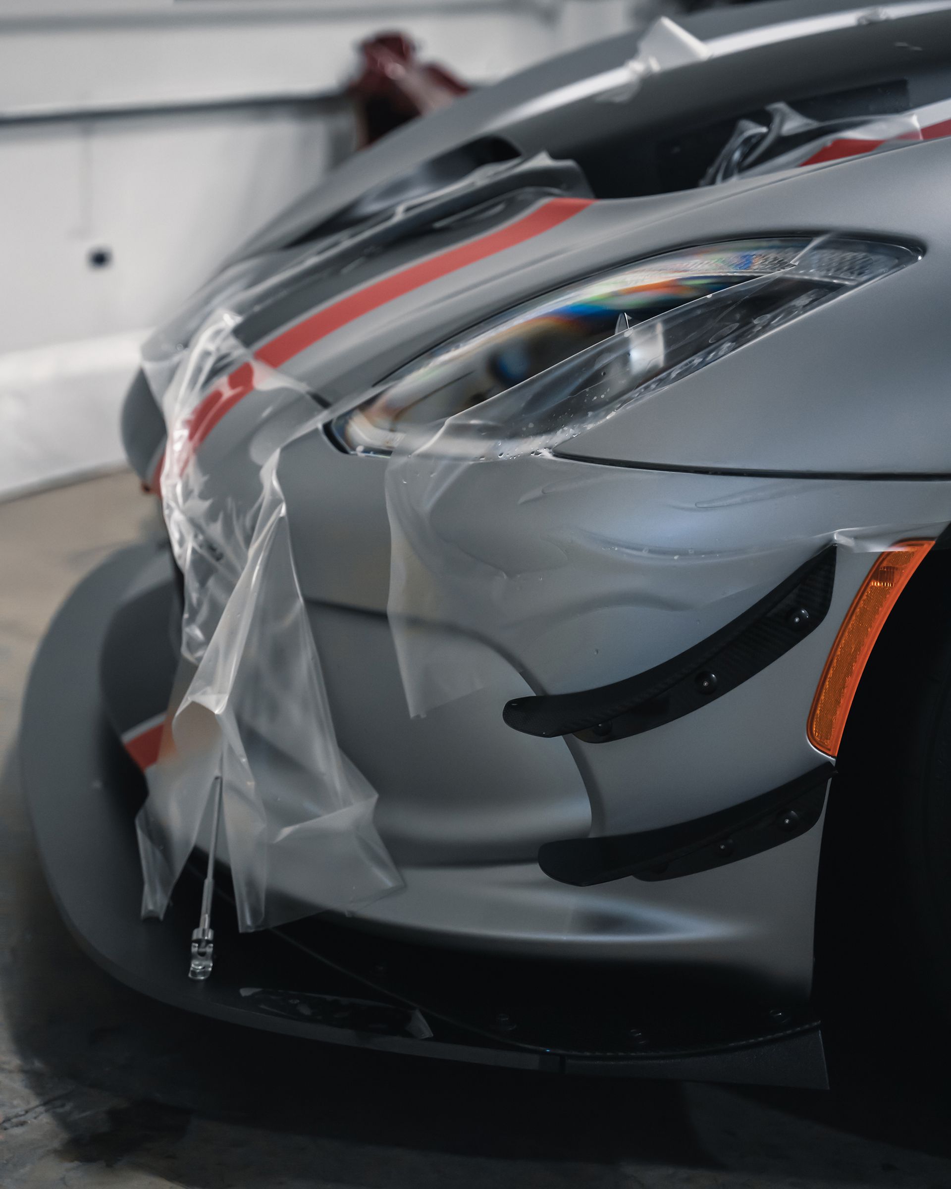 Silver sports car front with protective plastic, red stripes. Garage setting.
