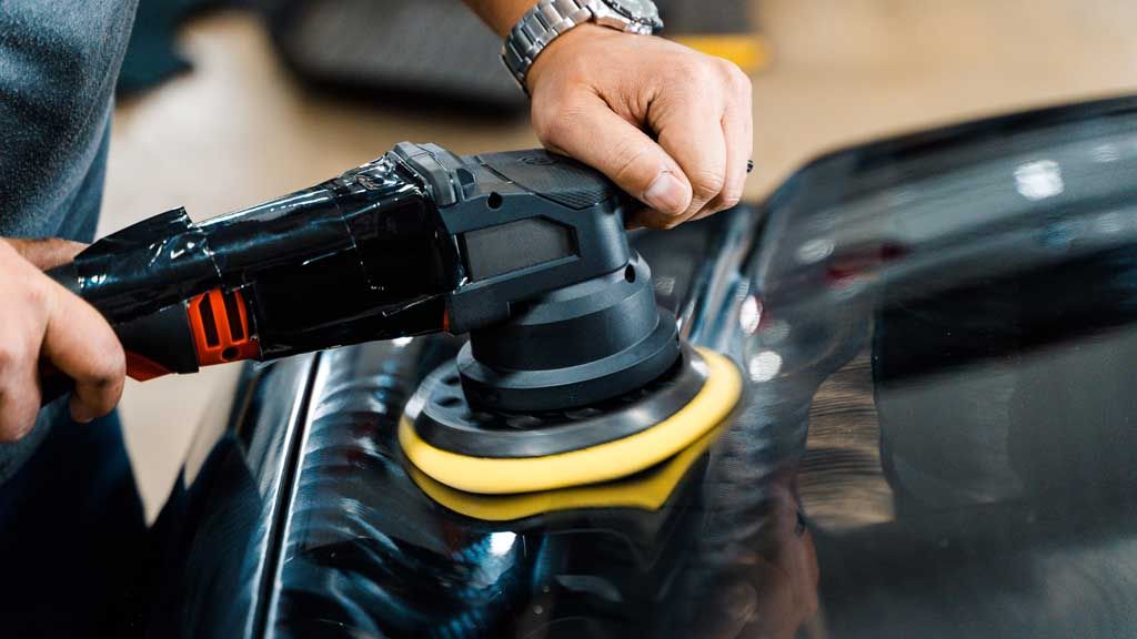 Person using an electric polisher on a black car panel.