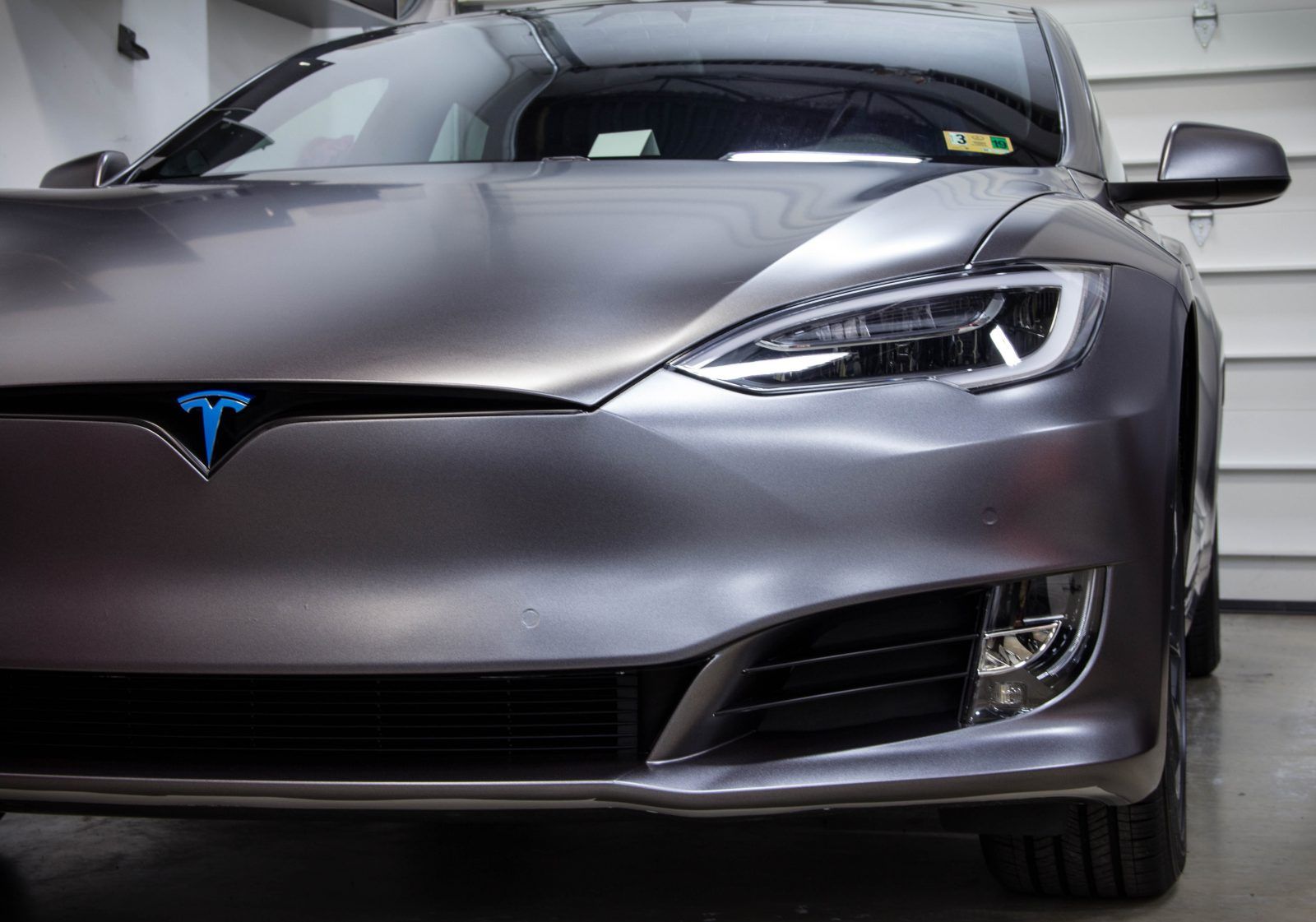 A grey Tesla Model S parked in a garage, front view. Features a blue Tesla logo.