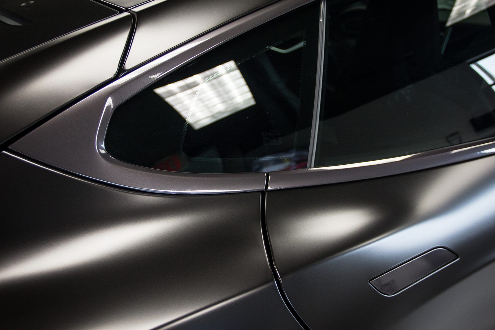 Close-up of a sleek, matte black car's side window and body panel; metallic trim reflects light.
