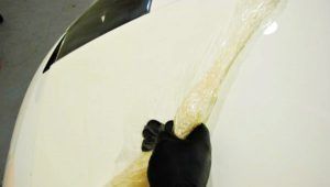 Gloved hand holding a long, wrapped object on a white car hood.