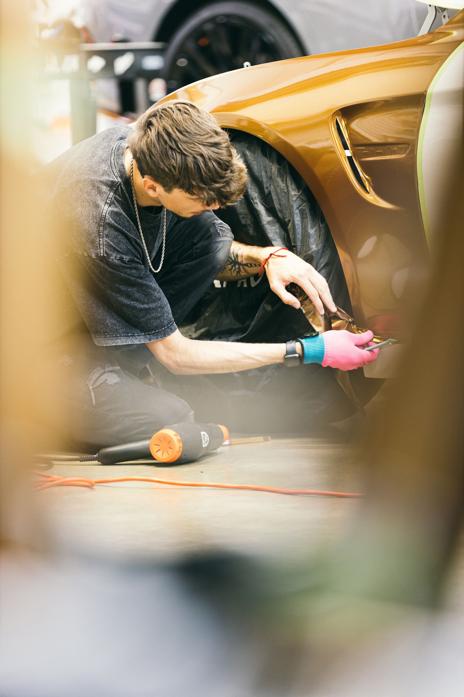 Person kneeling, working on the front fender of a gold car. Holding a tool, wearing pink gloves, black shirt, in a garage.