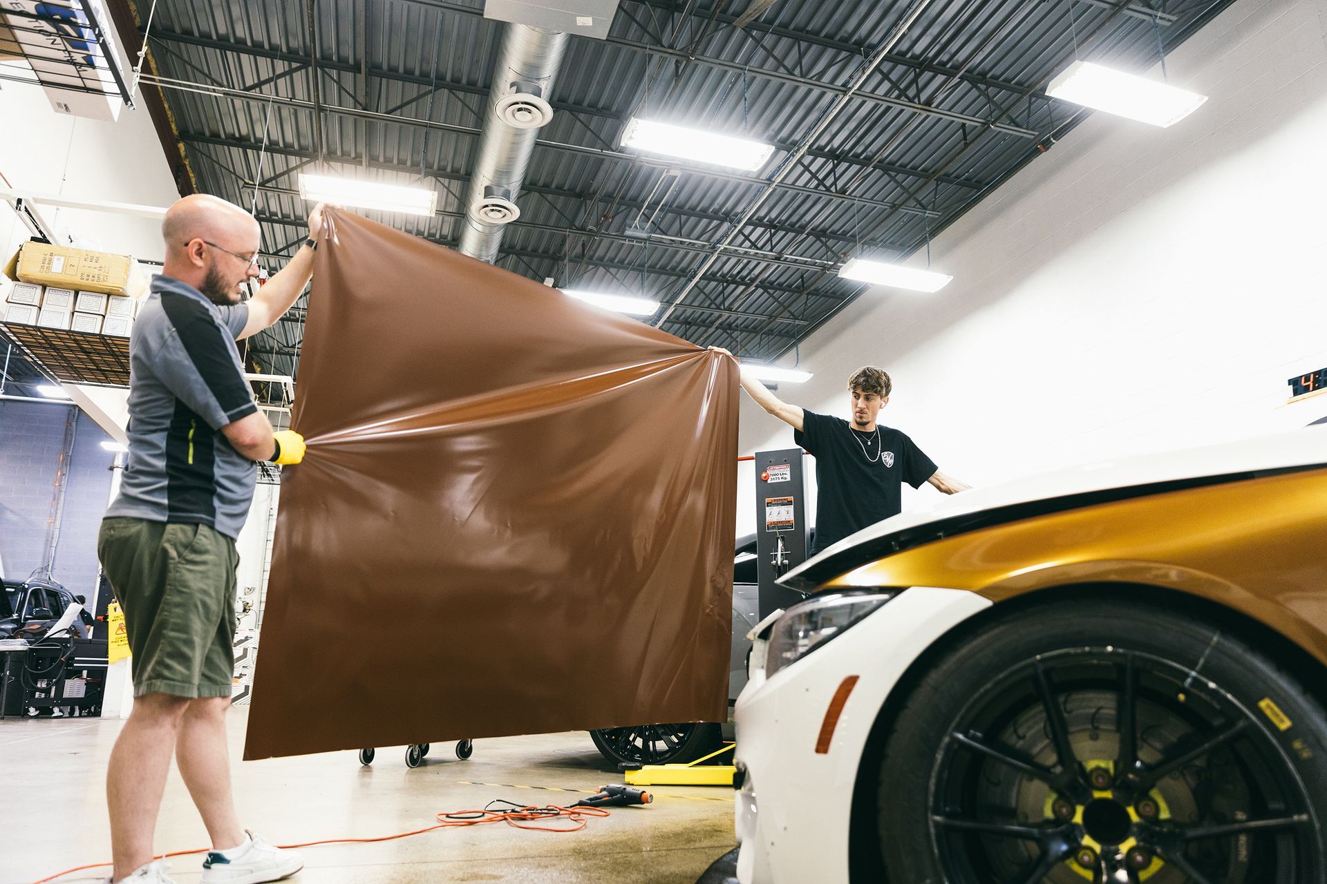 Two men applying brown vinyl wrap to a white and gold car in a garage.