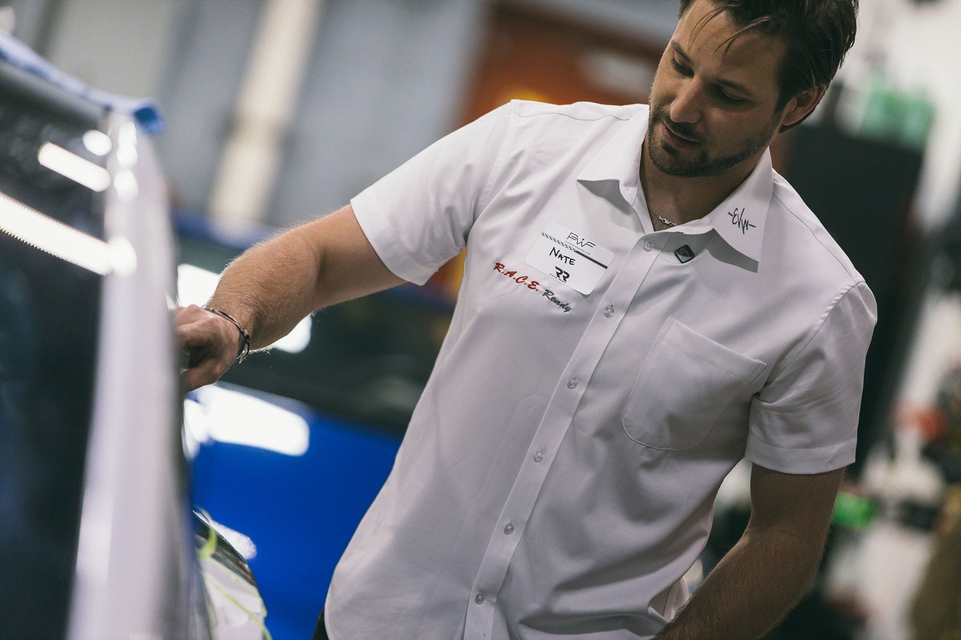 Man in white shirt working on car, possibly applying vinyl wrap. Indoors, shop setting.
