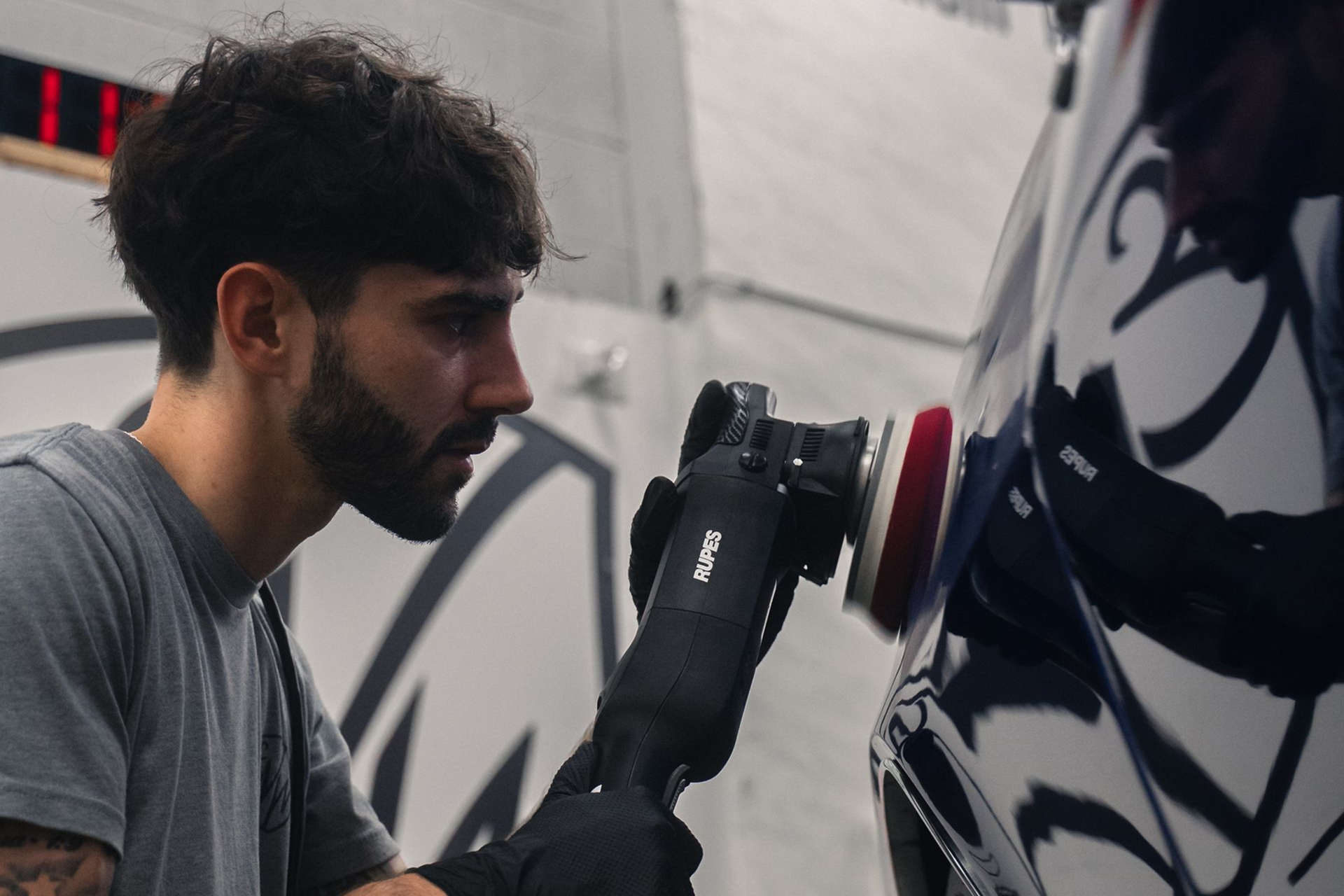 Man using a polisher on a car in a shop. He wears gloves, focused, and works on the car's side.