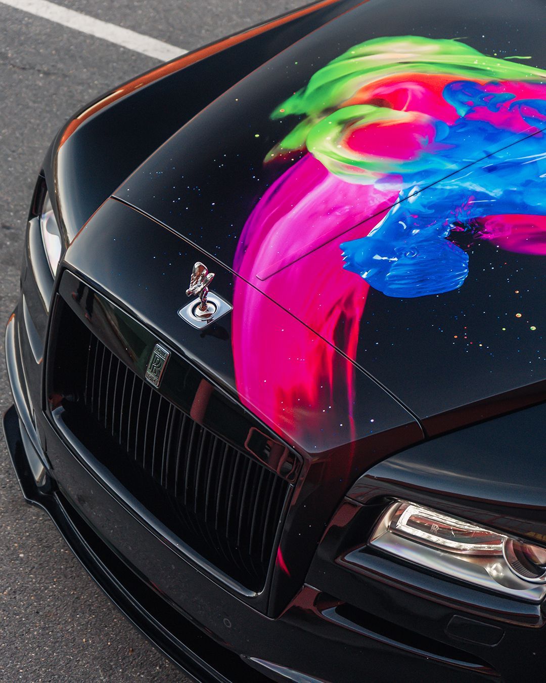 Black Rolls-Royce with colorful abstract paint on the hood, parked outside.