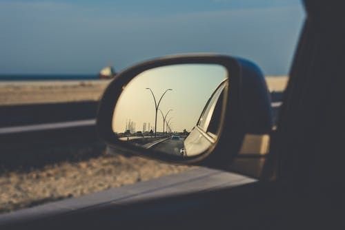 Side mirror reflecting a road with lamp posts, a car door, and a coastal setting.