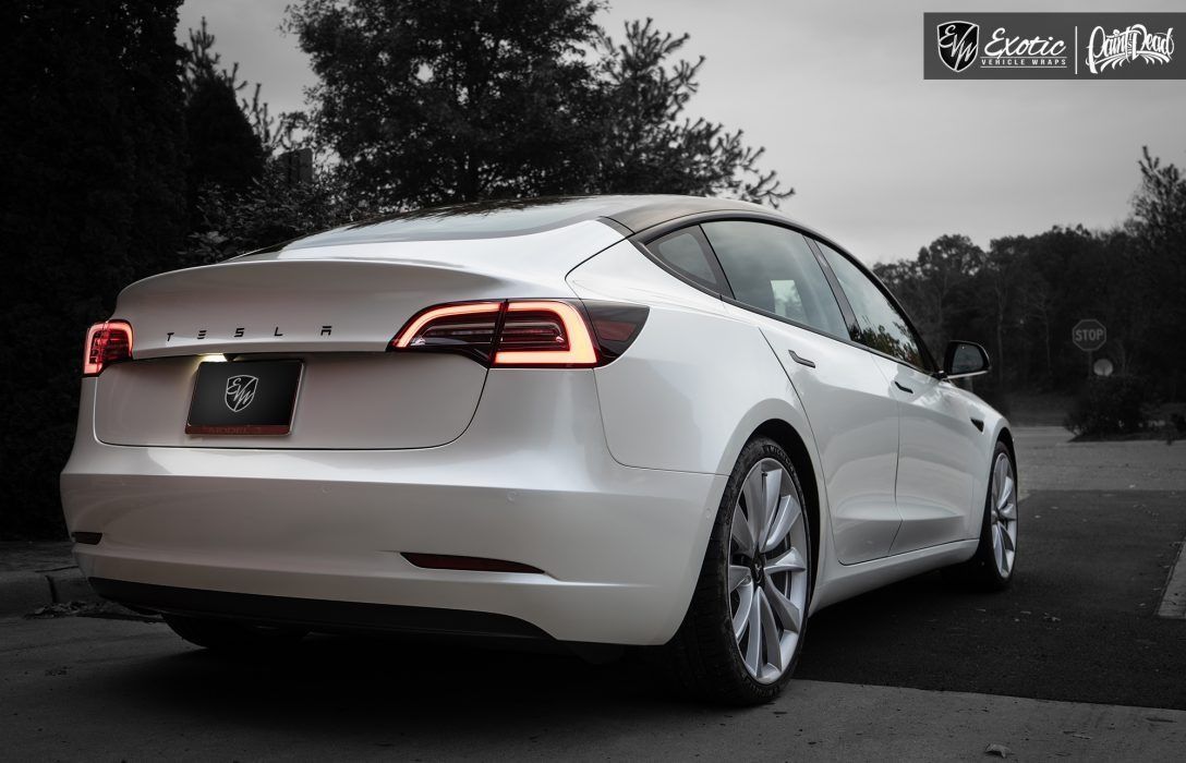 White Tesla sedan parked outside, rear view. Tail lights illuminated, silver wheels, grayscale background.