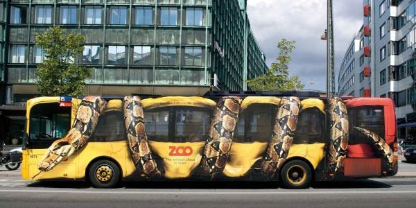 A yellow bus wrapped in a giant snake advertisement for the zoo, in a city setting.