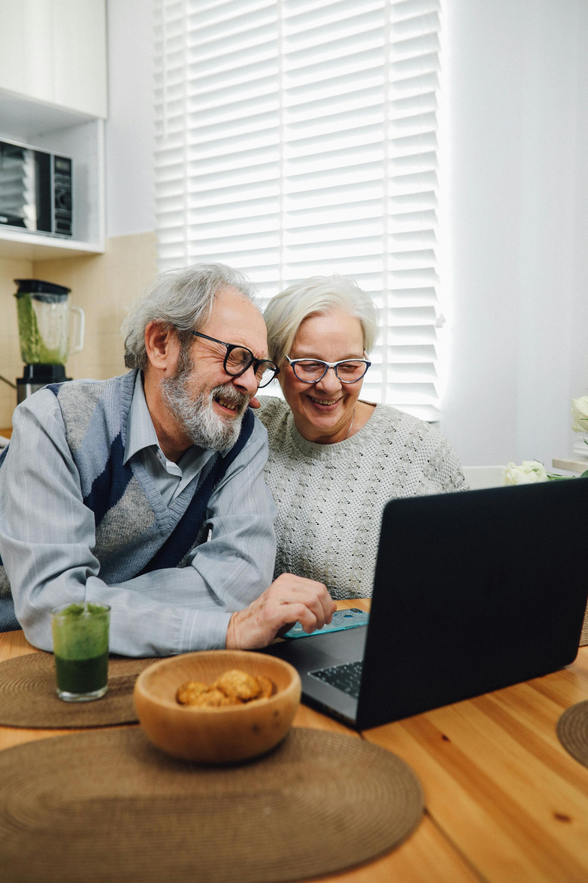 Elderly couple smiles, looking at laptop in kitchen. Cookies and green drink sit on table.