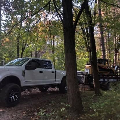 White pickup truck with a trailer carrying a CAT skid steer loader parked in a wooded area.