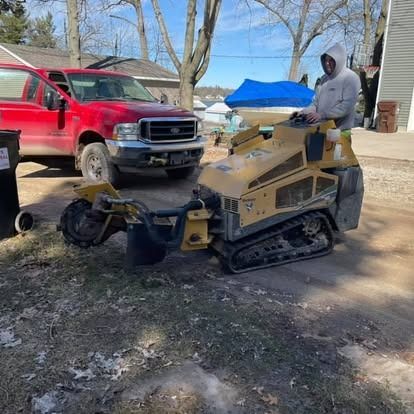 Man operating a yellow stump grinder next to a red pickup truck, in front of a house.