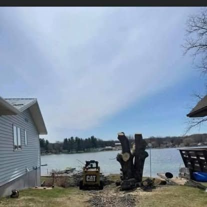 A stump next to a lake. A small Cat loader sits nearby. A house is on the left, another on the right.