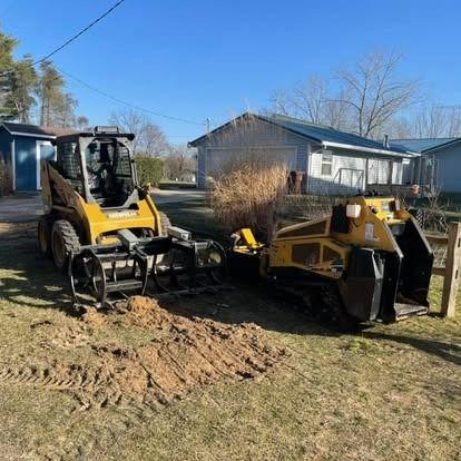 Two yellow construction vehicles on a grassy yard. One uses a grapple, the other a bucket. Sunny day.