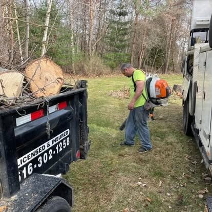 Man using a leaf blower, a trailer loaded with wood, and a truck on a grassy lawn.