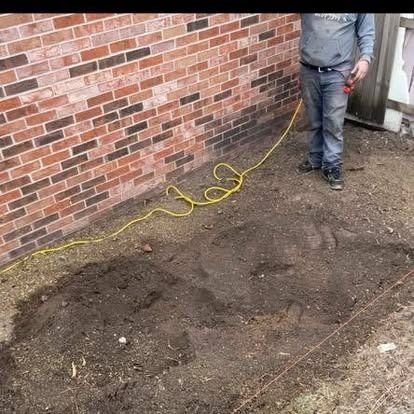 Man stands beside a brick wall, next to a dug-up patch of dirt, holding an electrical cord.