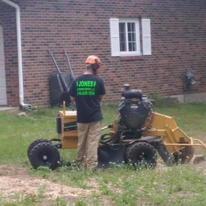 Man operating a yellow stump grinder in a yard near a brick building.