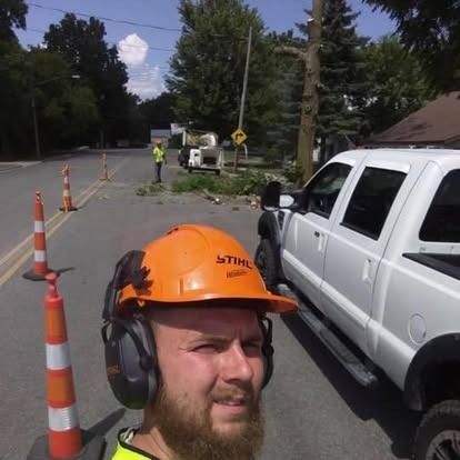 Man in safety gear, selfie-style. Roadside tree work: a white truck, cones, and workers are visible.