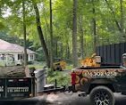 Trucks hauling wood and equipment, near a home and wooded area.