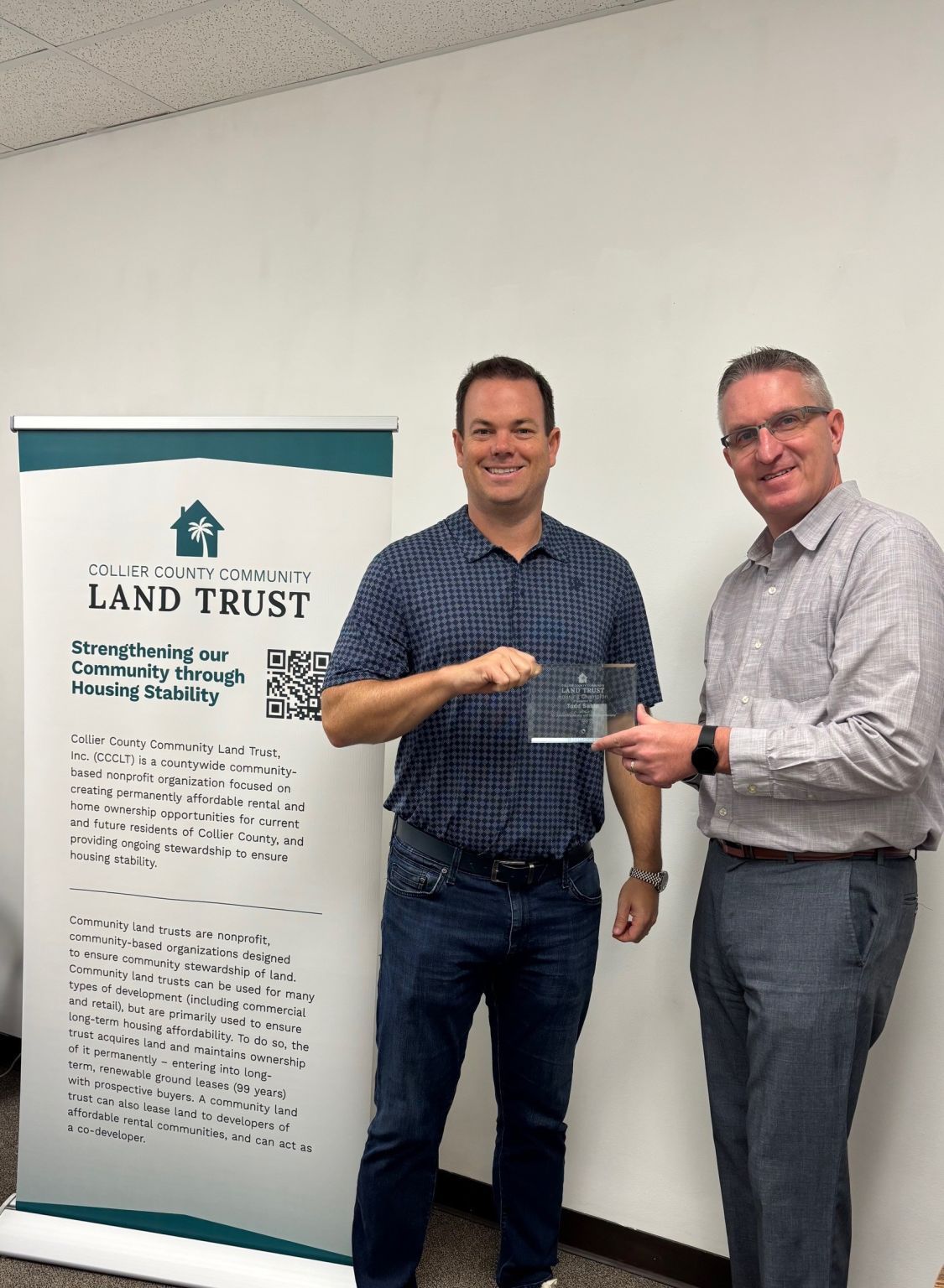 Two men holding an award; one points to a banner for a land trust. White wall background.
