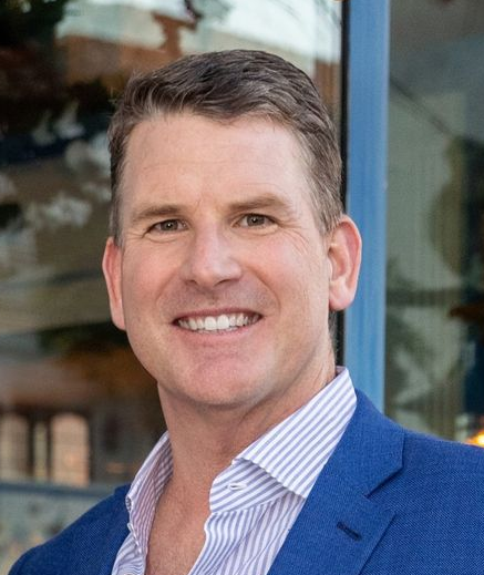 Sean. P. Murphy  in blue blazer smiling, light blue and white striped shirt, blurry outdoor setting.