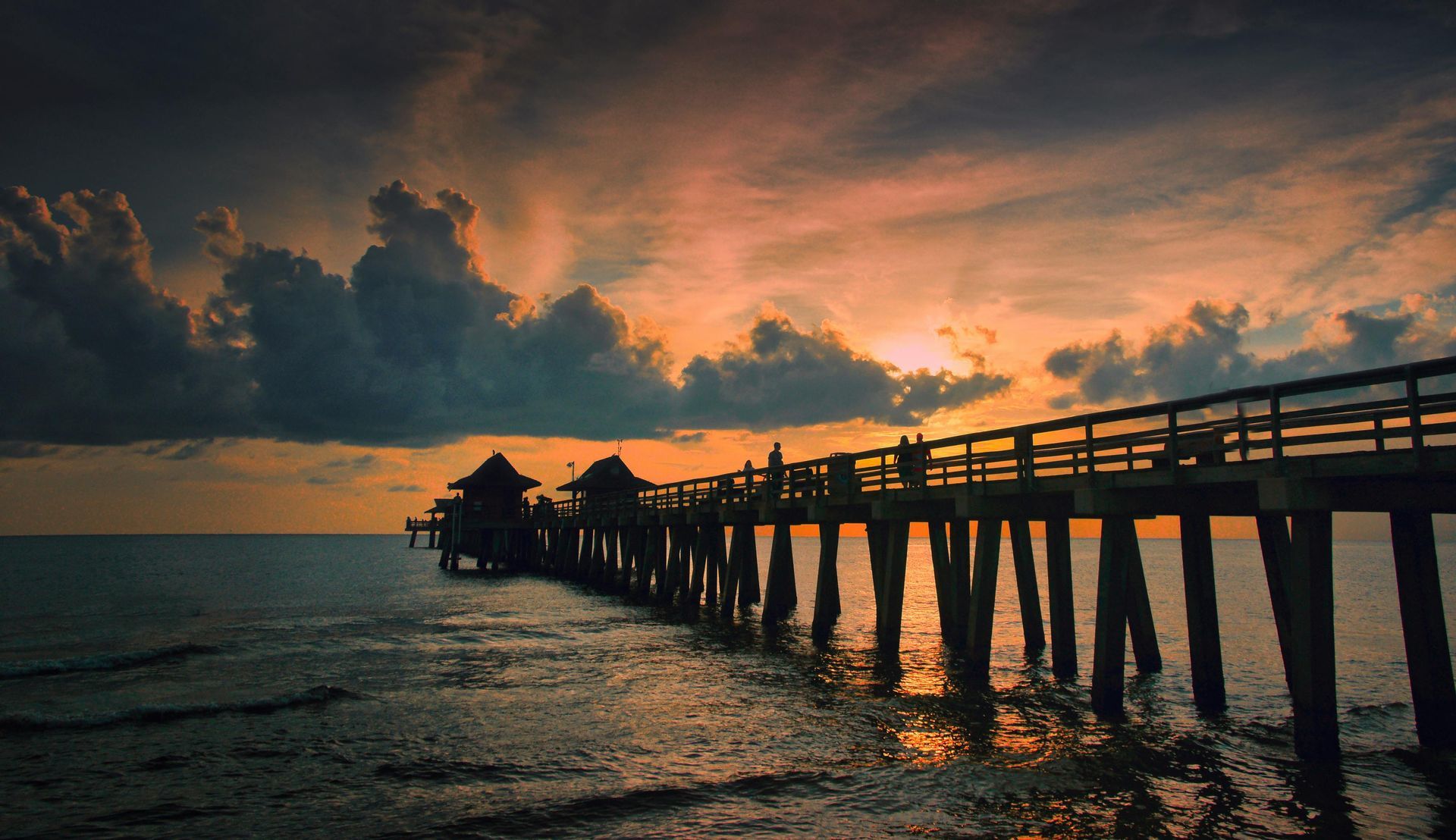 Sunset over the ocean. Pier with silhouetted figures against orange and dark gray sky.