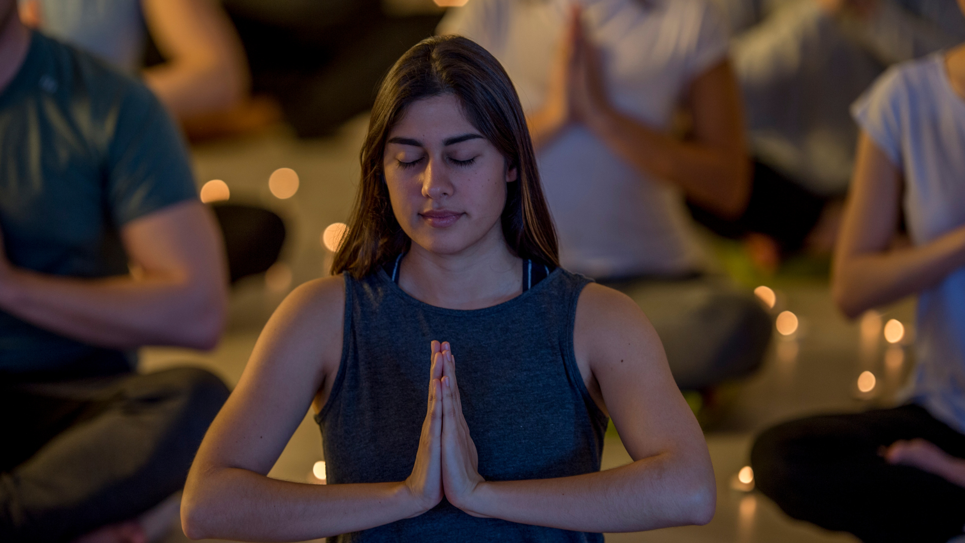 Woman in a yoga class, hands in prayer pose, eyes closed, in front of blurred people and candles.