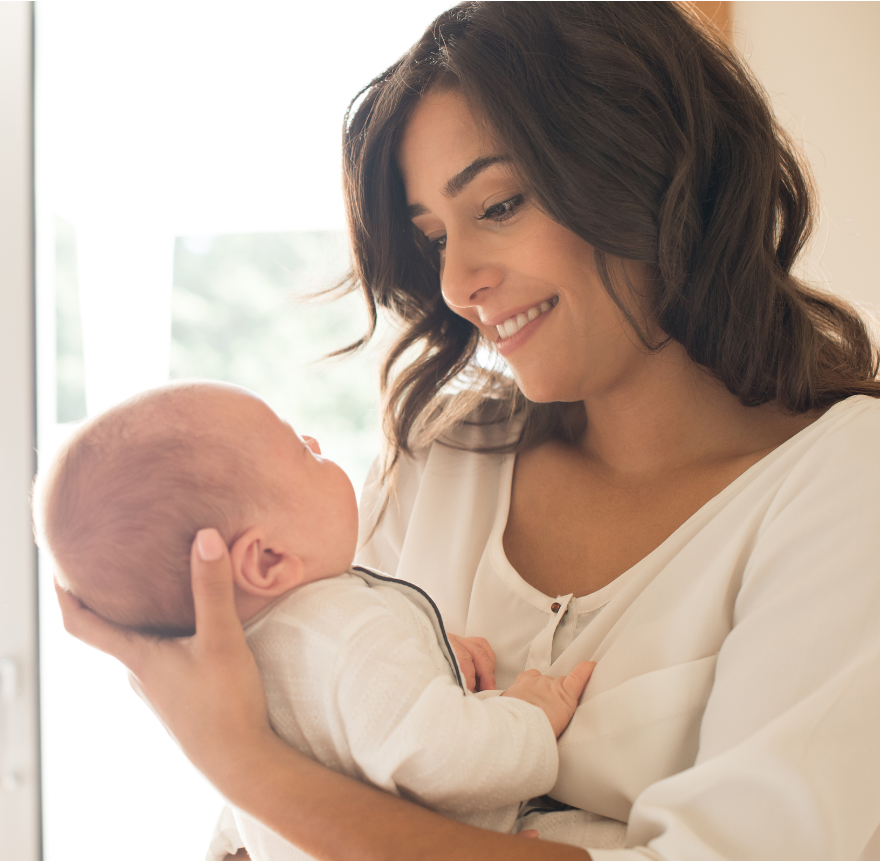 Woman smiles, cradling a baby in her arms, looking at it lovingly in a bright room.