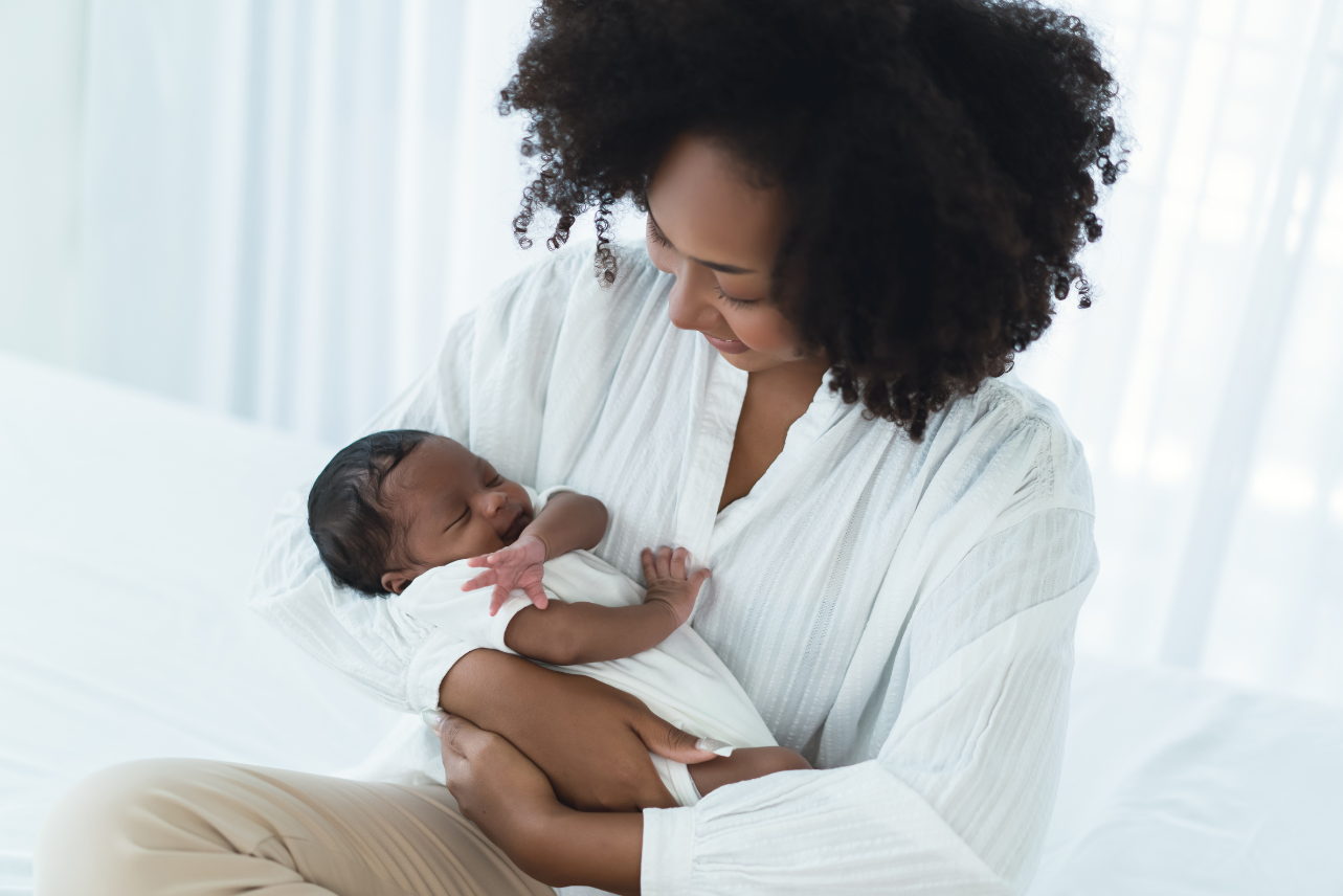 Woman with dark curly hair smiles down at newborn baby in white onesie.