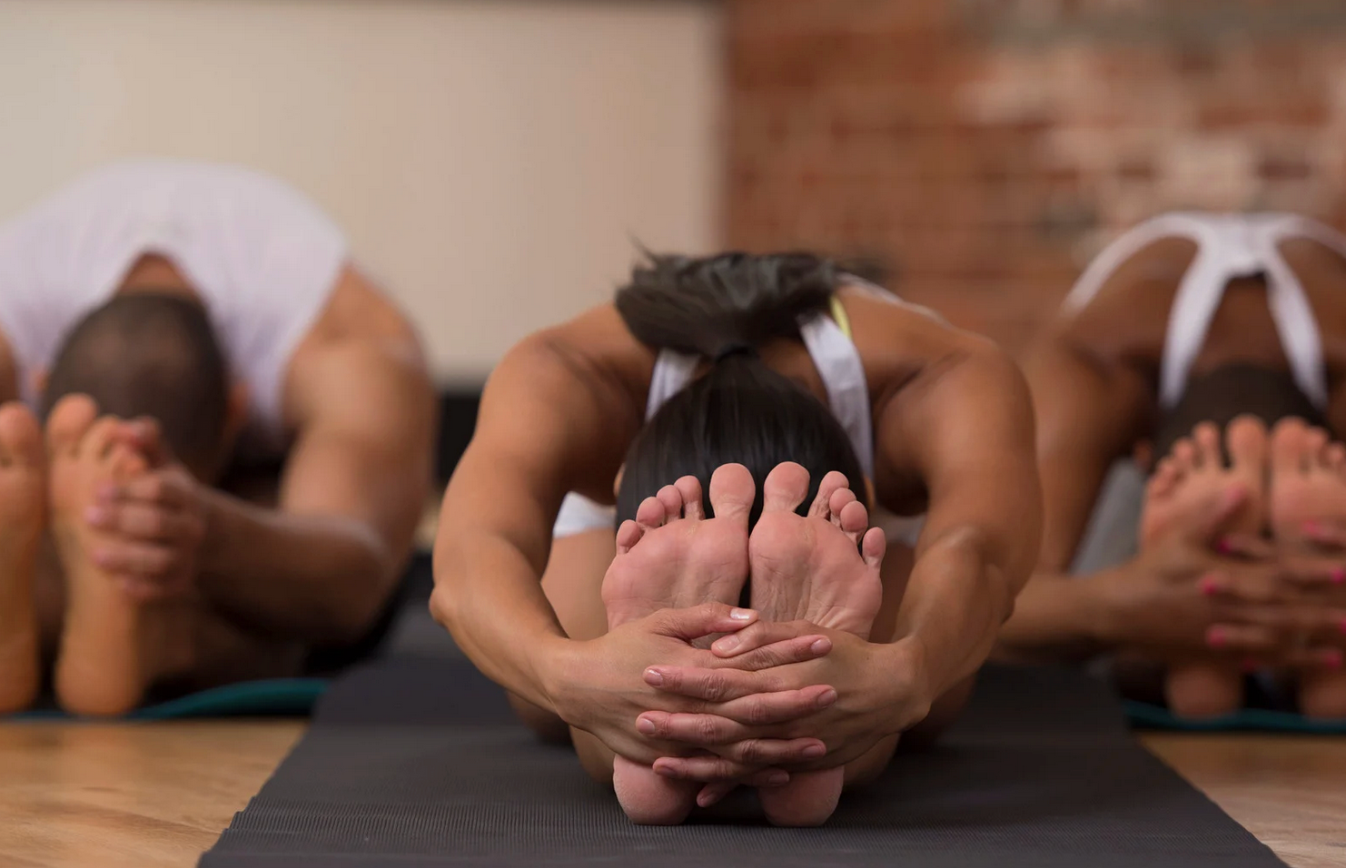 Three people in a yoga class, bending forward to touch toes, hands clasped around feet, on mats.