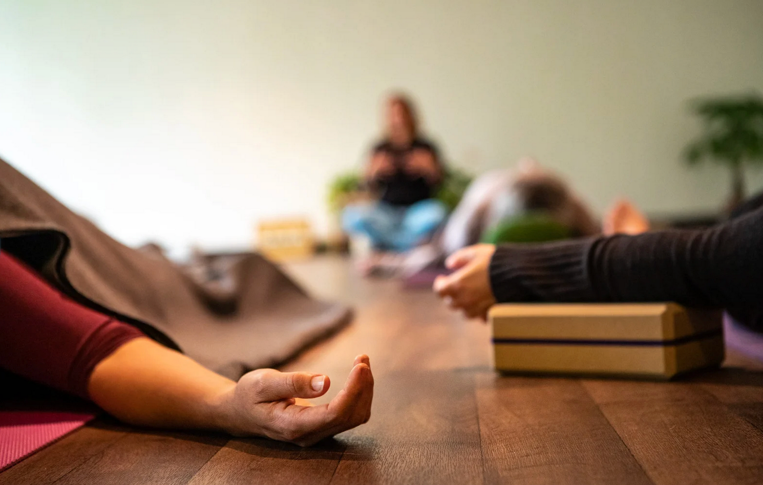 People in a yoga class lying on wooden floor, some with arms outstretched, using blocks.