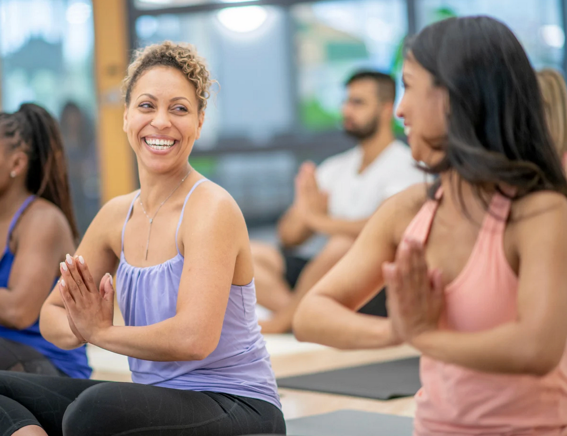 Yoga class: women smiling, hands in prayer pose, indoors with others.