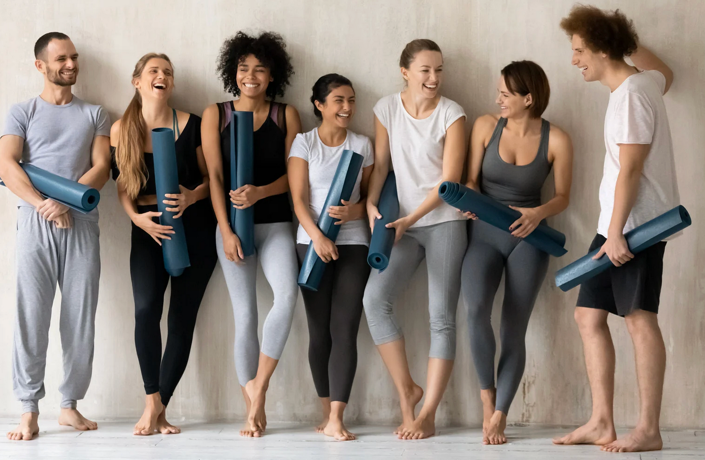 Group of people smiling, holding yoga mats, dressed in workout clothes, standing by a wall.