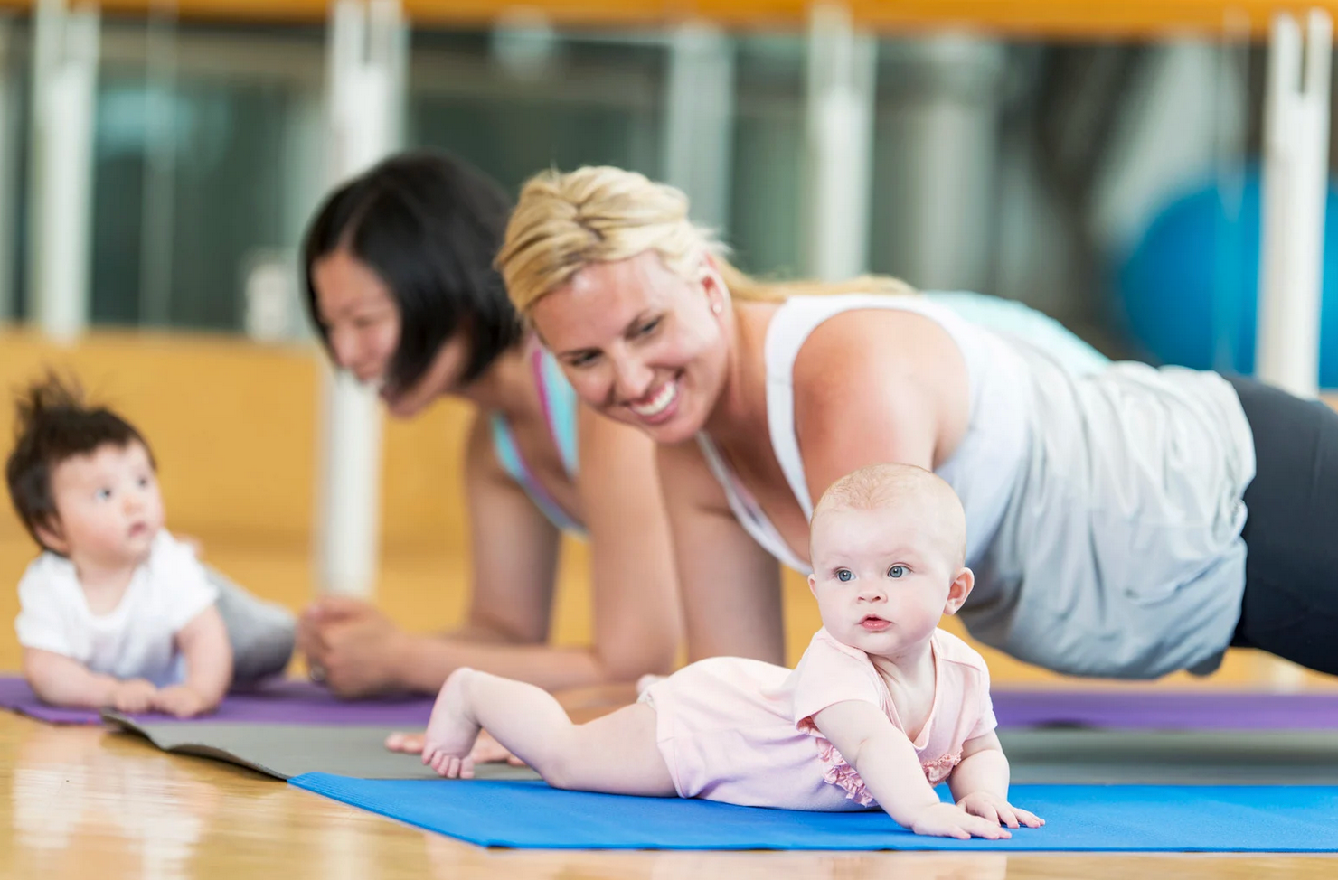 Mothers doing plank exercise with babies on mats in a gym. One baby looks at the camera.