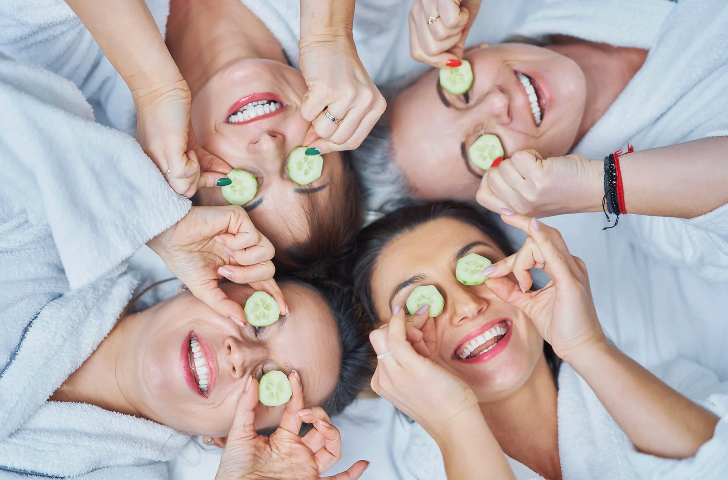 Four women in white robes at a spa, smiling with cucumber slices over their eyes.