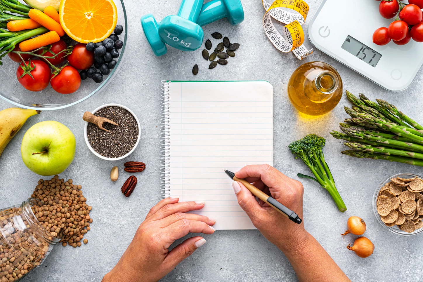 Person writing in notebook, surrounded by healthy food, weights, and a scale on a table.