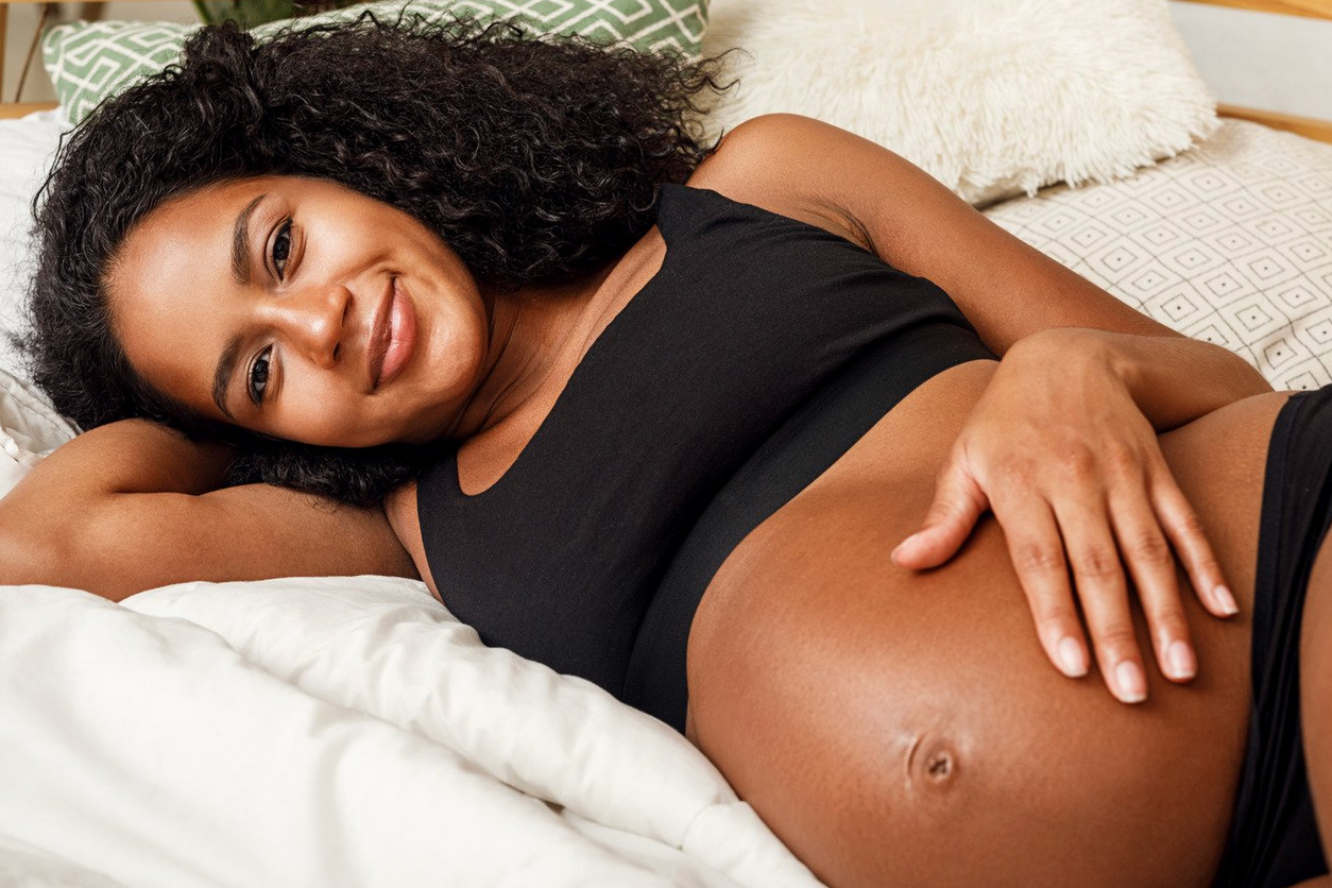 Pregnant woman with dark skin smiles, resting on a bed, hand on her belly.