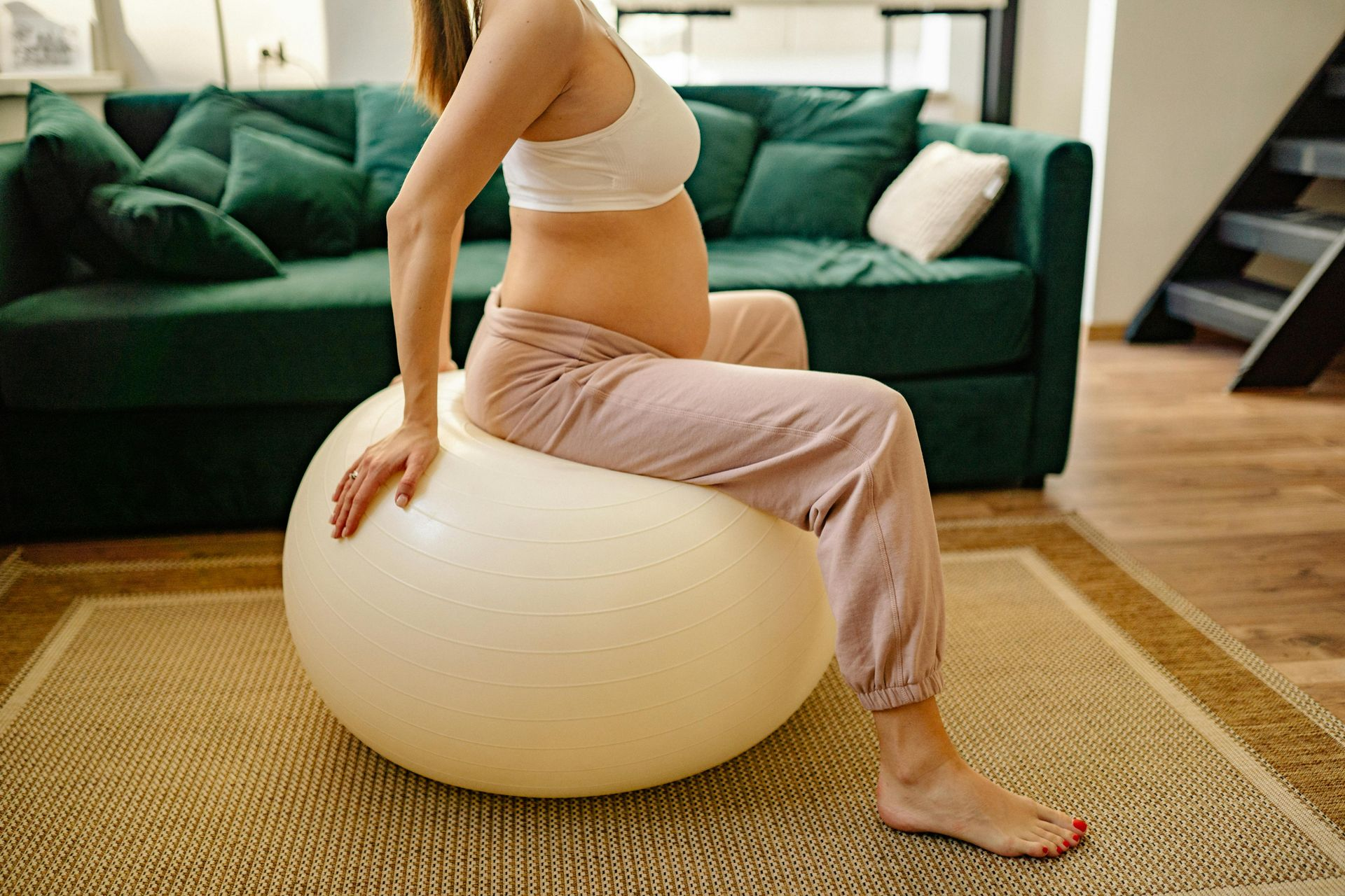 Pregnant woman sitting on a large exercise ball in a living room, stretching.